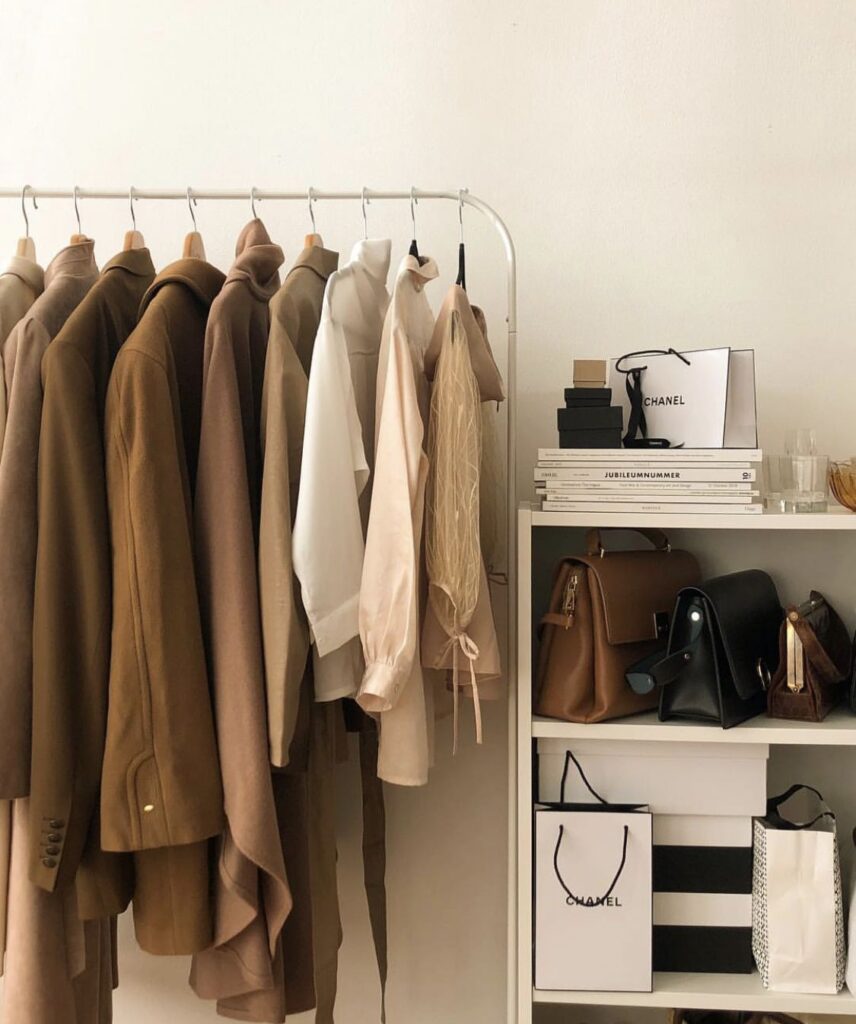 Minimalist wardrobe display with neutral-toned coats and blouses hanging on a white clothing rack beside a shelving unit holding designer handbags, books, and black-and-white Chanel shopping bags.