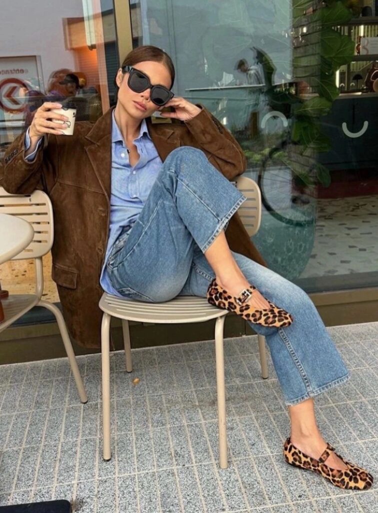 A woman sitting at an outdoor café wearing a brown oversized blazer, light blue button-down shirt, straight-leg jeans, and leopard print flats, holding a coffee cup and wearing black sunglasses.
