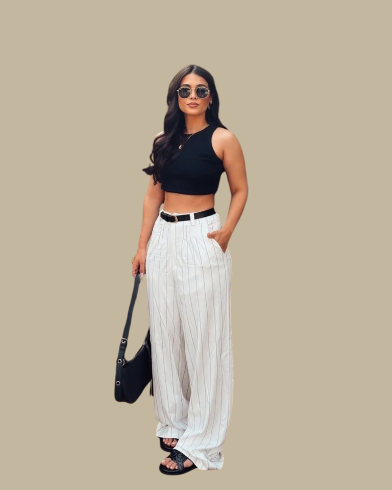 Woman wearing a black midi dress with a tank top and a flowing, full skirt, accentuated with a black and gold belt at the waist. She is holding sunglasses and carrying a small structured brown leather bag and white strappy sandals.
